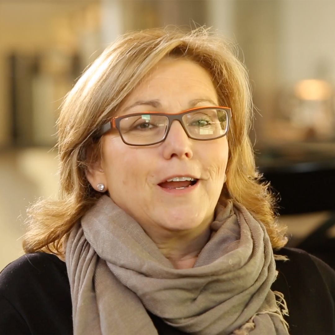 A woman with blonde hair and glasses smiles while sitting indoors. She is wearing a gray scarf and a black top, with a softly lit background showing blurred furniture.